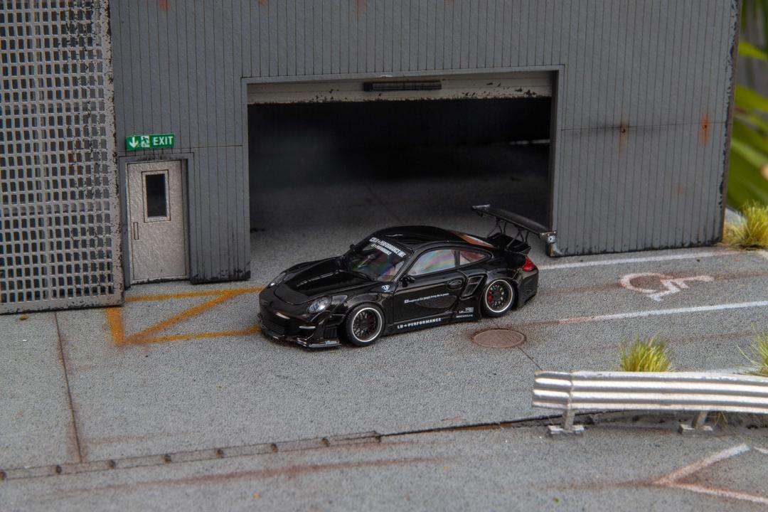 Star Models 1:64 Porsche 997 widebody in gloss black, front three-quarter view in a loading-dock diorama