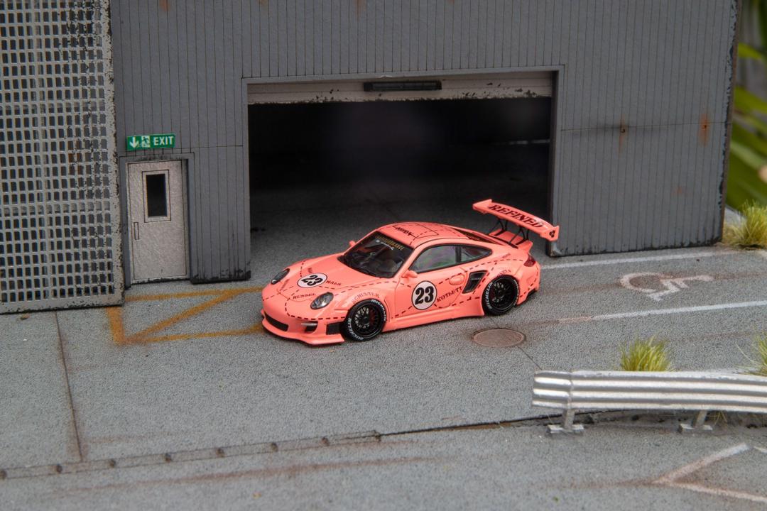 Star Models 1:64 Porsche 997 widebody in Pink Pig livery with number 23 roundel, front three-quarter view in a loading-dock diorama