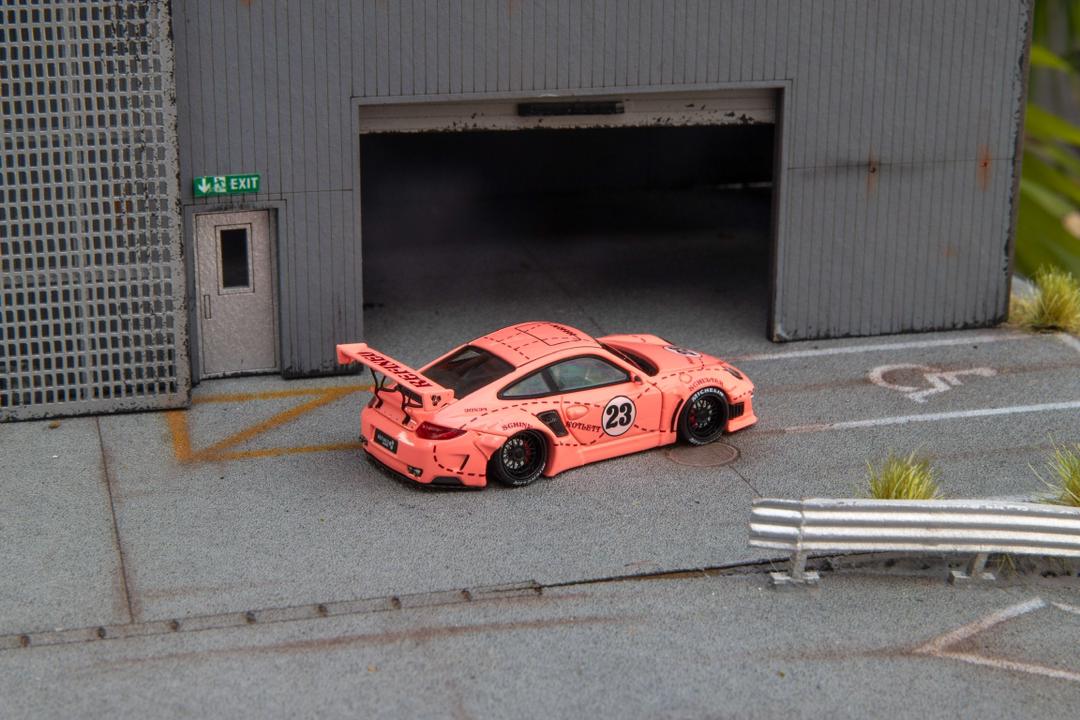 Star Models 1:64 Porsche 997 widebody in Pink Pig livery with number 23 roundel, rear three-quarter showing the large rear wing and butcher-cut graphics