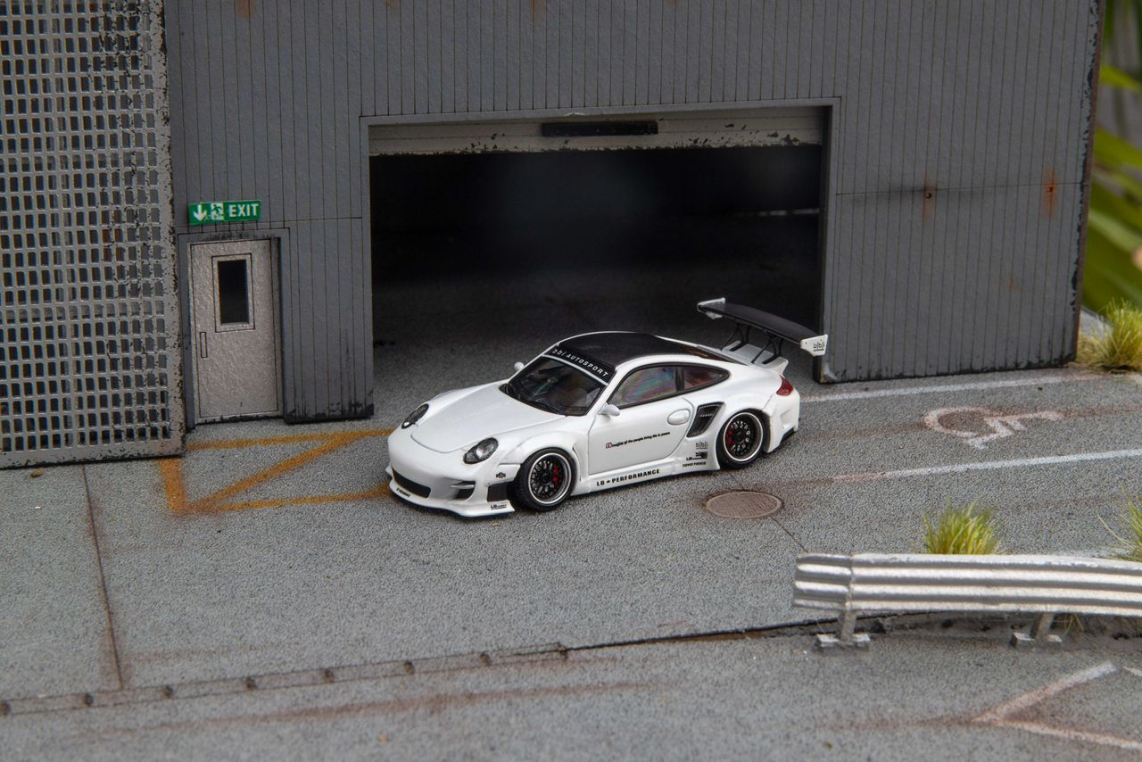 Star Models 1:64 Porsche 997 widebody in white with a carbon roof, front three-quarter view in a loading-dock diorama